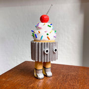 Wooden sculpture of a cupcake with white frosting, sprinkles, and a cherry on top. Its wrapped in a silver wrapper and stands on 2 legs, with a pair of eyes as its only facial feature.