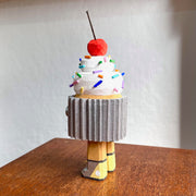 Wooden sculpture of a cupcake with white frosting, sprinkles, and a cherry on top. Its wrapped in a silver wrapper and stands on 2 legs, with a pair of eyes as its only facial feature.