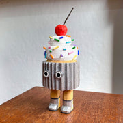 Wooden sculpture of a cupcake with white frosting, sprinkles, and a cherry on top. Its wrapped in a silver wrapper and stands on 2 legs, with a pair of eyes as its only facial feature.