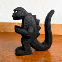 Whittled wooden sculpture of a black Godzilla character, with white eyes and a stern expression. It stands with its arms slightly raised at its sides and its large tail behind it.
