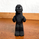 Whittled wooden sculpture of a black Godzilla character, with white eyes and a stern expression. It stands with its arms slightly raised at its sides and its large tail behind it.