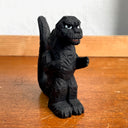 Whittled wooden sculpture of a black Godzilla character, with white eyes and a stern expression. It stands with its arms slightly raised at its sides and its large tail behind it.