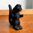 Whittled wooden sculpture of a black Godzilla character, with white eyes and a stern expression. It stands with its arms slightly raised at its sides and its large tail behind it.
