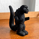 Whittled wooden sculpture of a black Godzilla character, with white eyes and a stern expression. It stands with its arms slightly raised at its sides and its large tail behind it.