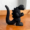 Whittled wooden sculpture of a black Godzilla character, with white eyes and a stern expression. It stands with its arms slightly raised at its sides and its large tail behind it.