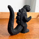 Whittled wooden sculpture of a black Godzilla character, with white eyes and a stern expression. It stands with its arms slightly raised at its sides and its large tail behind it.