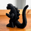 Whittled wooden sculpture of a black Godzilla character, with white eyes and a stern expression. It stands with its arms slightly raised at its sides and its large tail behind it.
