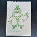 A green line-drawn cartoon character on a textured beige background, with the text 'DOING WHAT I CAN.' at the bottom, photographed against a dark textured surface.