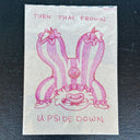 A  pink hand-drawn illustration of a crown on a beige background, with the words 'TURN THAT FROWN' at the top and 'UPSIDE DOWN' at the bottom, photographed on a dark textured surface.