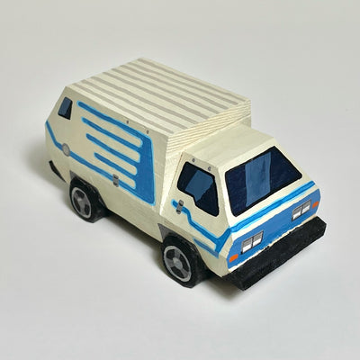 Carved and assembled wooden sculpture of a VW Bus, cream colored with blue patterned line accents and an overall bulky body.