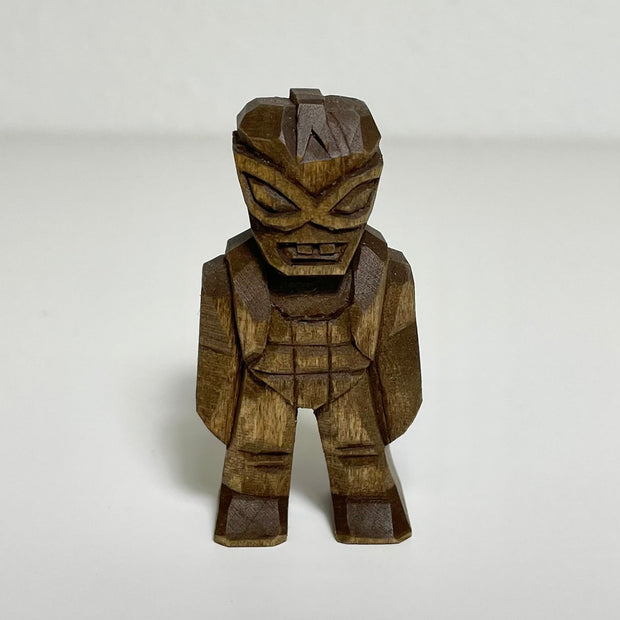 Carved, unpainted wooden sculpture of an alien with an oversized head and buck teeth. It sports goggles and other armor like clothing.
