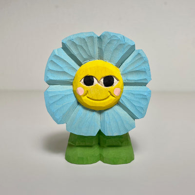 Carved and painted wooden sculpture of a smiling blue flower with a yellow face. It stands on 2 green feet, with no arms or body.