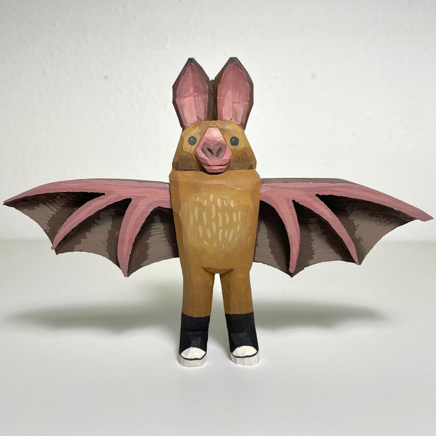 Carved and painted wooden sculpture of a brown bat with pink veined wings spread out and pink ears atop its head. It wears a pair of black sneakers.