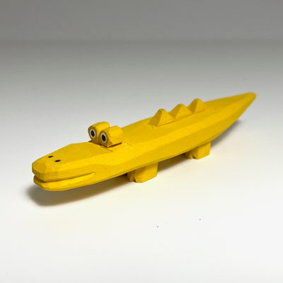 Carved and painted wooden sculpture of a yellow alligator, its body resembling a flat banana with spikes on the back. It has a pair of eyes atop its head and a long closed mouth snout.