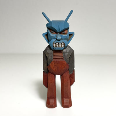 Carved and painted wooden sculpture of an alien with an angry blue face and red eyes. Its teeth are gritted and its body wears brown and grey armor suit.