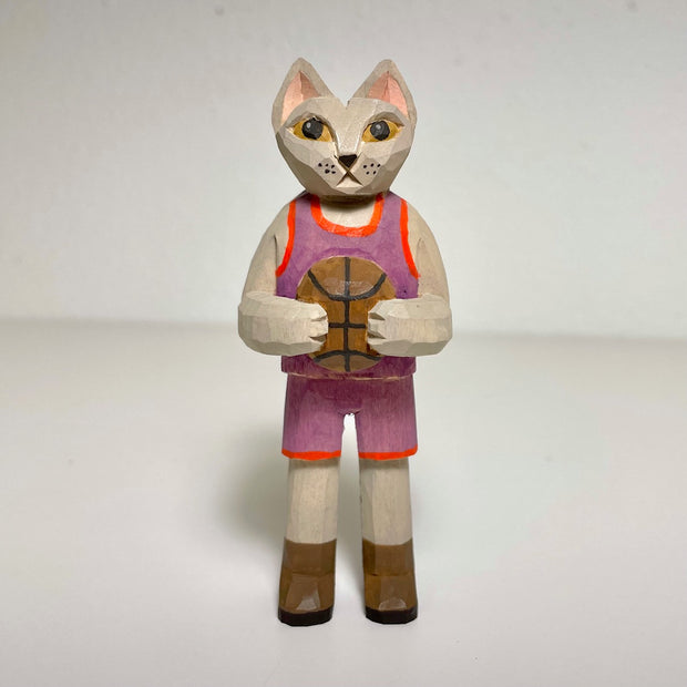 Carved and painted wooden sculpture of a tan cat wearing a basketball uniform, purple with red lining. It holds a basketball to its chest and wears brown boots.