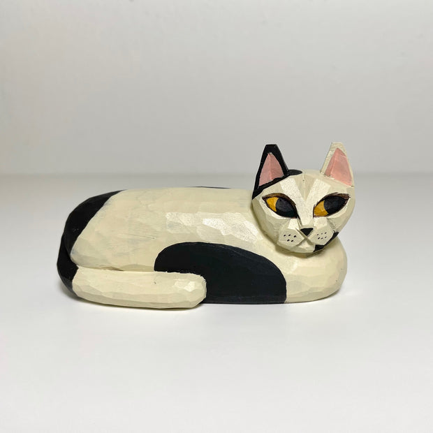Carved and painted wooden sculpture of a white cat with black spots laying down, folded into itself with its head raised up.
