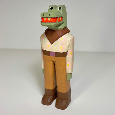 Carved and painted wooden sculpture of a crocodile wearing brown pants and a collared floral patterned shirt. It wears a brown belt with matching brown boots and a neck bandana.