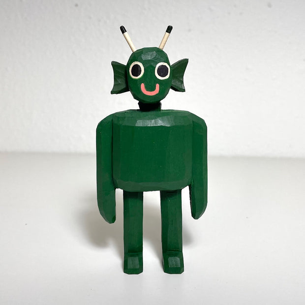 Carved and painted wooden sculpture of a green alien, with a face akin to the Creature from the Black Lagoon but with a simple sweet smile. It stands with its arms to its side.