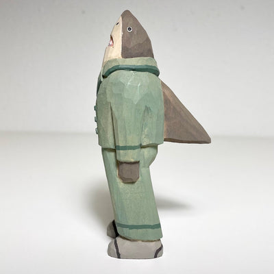 Carved and painted wooden sculpture of a shark standing on 2 legs and wearing a set of light green pajamas. It has a large back fin, which extends out of its pajamas.