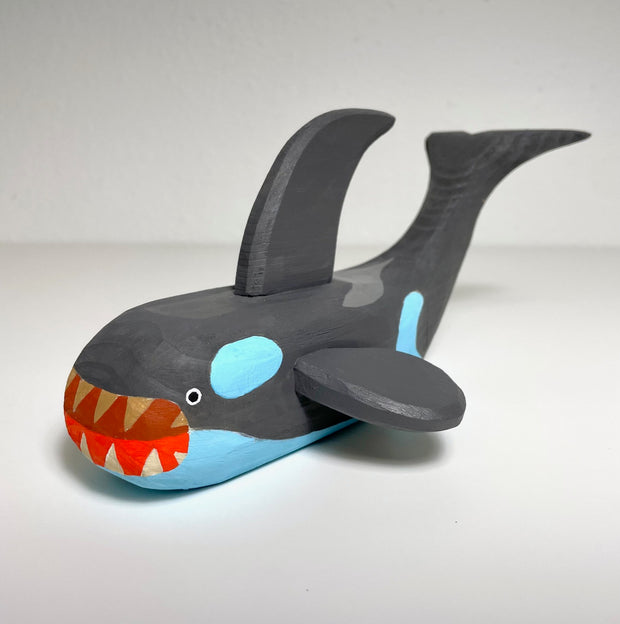 Carved and painted wooden sculpture of an orca whale, dark gray with blue coloring where is normally white. It has a large open red mouth revealing sharp teeth. Its positioned on its stomach with its tail raised, a large back fin and its side fins extended.