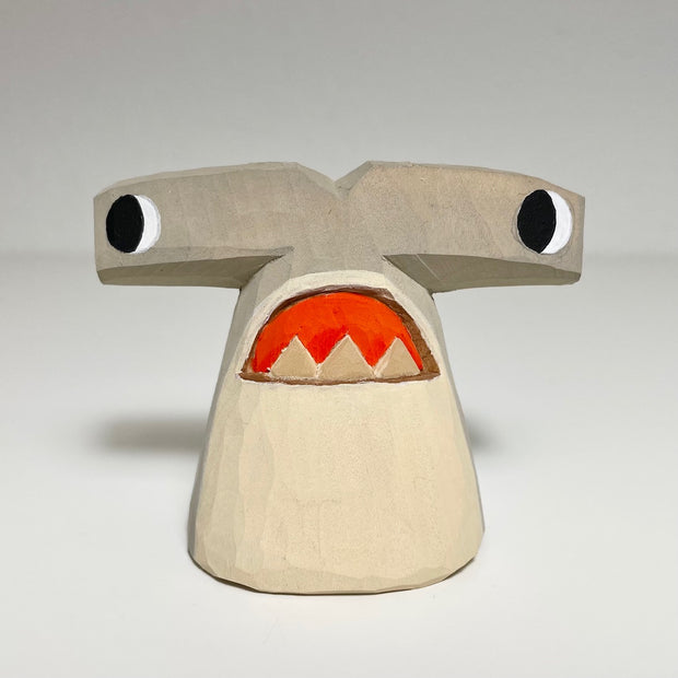 Carved and painted wooden sculpture of a hammerhead shark with a cartoon like expression, eyes looking off to the side and a downturned open mouth. 