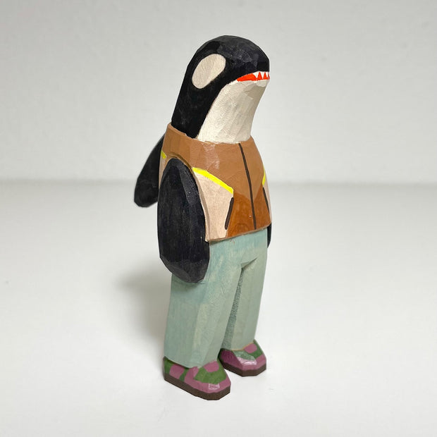 Carved and painted wooden sculpture of an orca, standing up on 2 legs like a person. It wears a brown vest, light blue pants and purple and green sneakers.