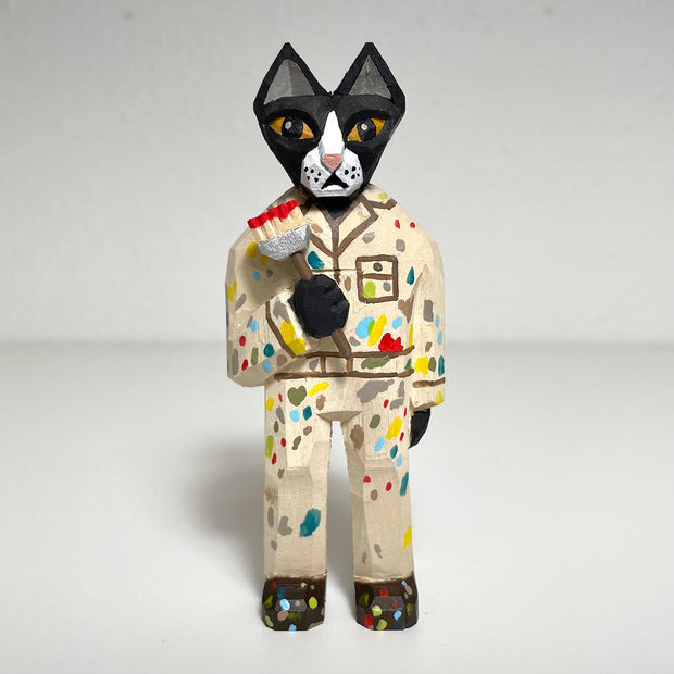 Carved and painted wooden sculpture of a black cat standing on 2 legs and wearing a cream colored painters outfit, and holding a paintbrush in one hand. Its jumper is covered in multicolor paint splotches.