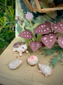 Diorama made of clay and paper of a character napping in a wooden structure in a tree, alongside a napping raccoon. Around them are flowers and a tiny hedgehog family with 3 babies.