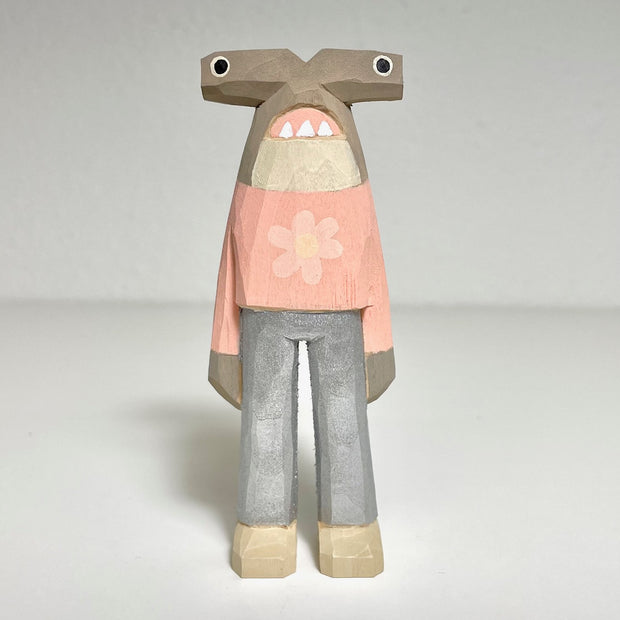 Carved and painted wooden sculpture of a hammerhead shark, standing on 2 legs like a person and wearing a pink shirt and silver pants. Its pink shirt has a flower on it and the shark has a cartoonish expression on its face.