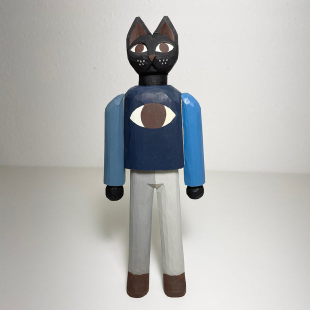 Carved and painted sculpture of a black cat standing on 2 legs, like a person. The cat has wide brown eyes and wears a blue raglan shirt with a large brown eye as the center design.