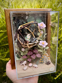 Diorama made of clay and paper of a character napping in a wooden structure in a tree, alongside a napping raccoon. Around them are flowers and a tiny hedgehog family with 3 babies.