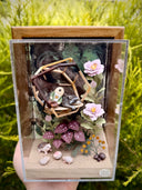 Diorama made of clay and paper of a character napping in a wooden structure in a tree, alongside a napping raccoon. Around them are flowers and a tiny hedgehog family with 3 babies.