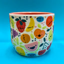 Ceramic cup covered with cute, cartoon illustrations of very colorful fruit with simple cartoon faces. Fruits include: grapes, cherries, citrus, pineapple, watermelon, etc.