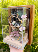 Diorama made of clay and paper of a character napping in a wooden structure in a tree, alongside a napping raccoon. Around them are flowers and a tiny hedgehog family with 3 babies.
