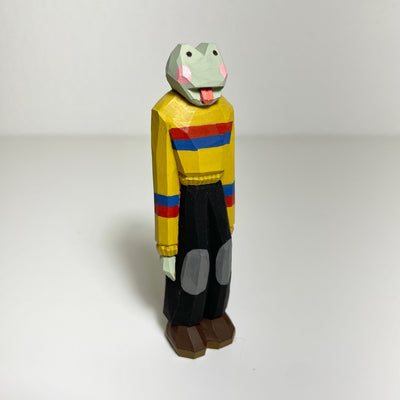 Carved and painted wooden sculpture of a frog, with its tongue out, standing on 2 legs like a person. It wears a yellow sweater with red and blue stripes and a pair of black pants with patch knees.