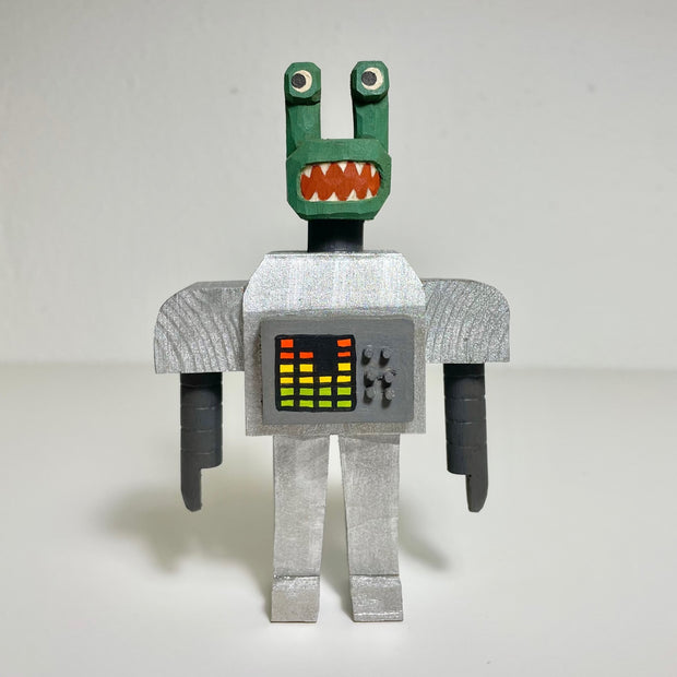 Carved and painted wooden sculpture of a green alien, its body composed of a boxy silver space outfit. On its body are controls and a colorful graph.
