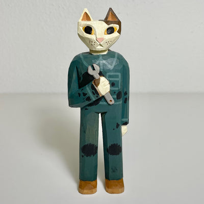 Carved and painted wooden sculpture of a calico cat, standing on 2 legs like a person. It wears a blue mechanic's jumpsuit and holds a wrench in its hand and has oil stains on its clothes.