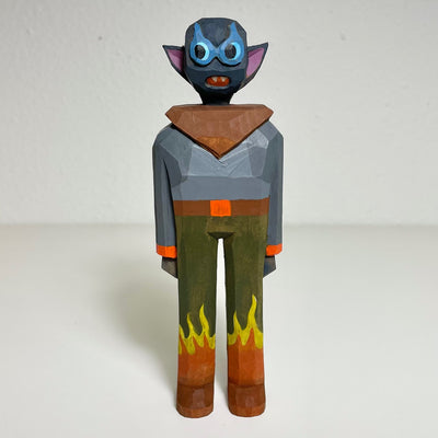 Carved and painted wooden sculpture of a funny faced alien, wearing a silver shirt with a bold brown triangle collar and olive green pants with flames at the bottom.