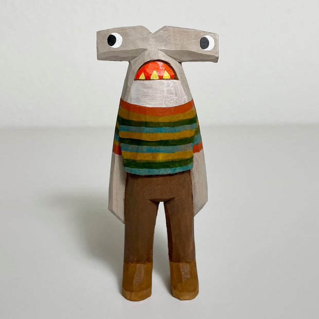 Carved and painted wooden sculpture of a hammerhead shark, standing on 2 legs like a person. Its facial expression is comical as it looks off to the side with a downturned open mouth. It wears a muted tone striped shirt and brown pants. 
