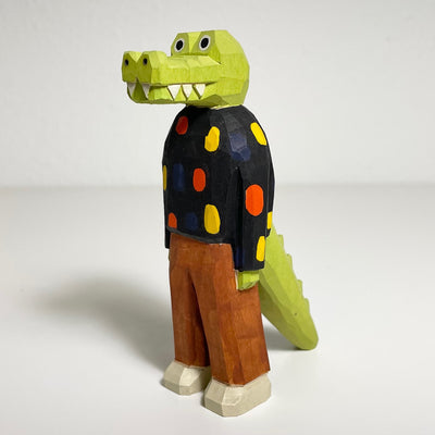 Carved and painted wooden sculpture of a light green gator standing on 2 legs, like a person. It wears a black sweater with multicolor polka dots and brown pants.
