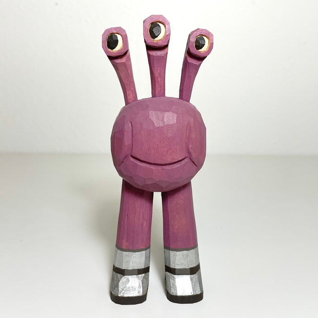 Carved and painted wooden sculpture of a a round bodied purple alien, with 3 eyes sticking out of its head. It has legs but no arms and wears shiny silver shoes.