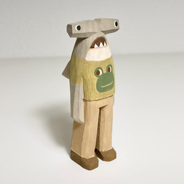 Carved and painted wooden sculpture of a hammerhead shark standing on 2 legs, like a person. It wears a olive green shirt with a graphic of a smiling frog, khaki pants and brown shoes.