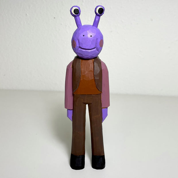Carved and painted wooden sculpture of a purple alien with a body like a person. It has a round head with eyes extended out like a snail. It wears an all brown ensemble, pants with a vest and shirt.