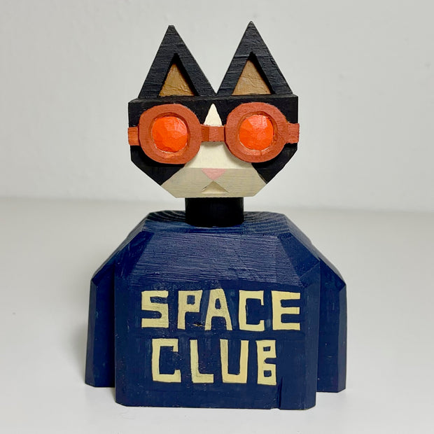 Carved and painted wooden sculpture of the bust of a black and white cat. It has round orange goggles and wears a navy shirt that reads "Space Club" in light yellow font.
