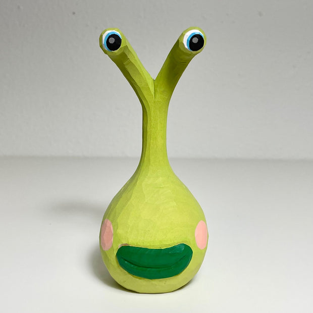Carved and painted wooden sculpture of a green plant alien's head. It's main head is rounded with dark green lips and pink cheeks and it has long extensions, like a snail, that lead to its eyes. 