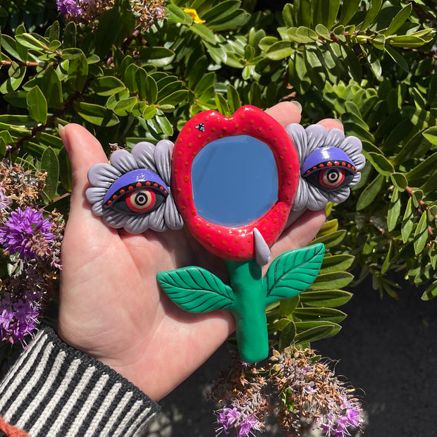 Polymer sculpture around a small mirror of a set of red lips, attached to a green plant stem. On either side of the lips are purple eyes embedded in flowers. A single fly rests on the top of the lips and a water droplet runs down the lower lip.