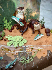 Diorama sculpture of 2 brown otters, one holds a fish in its hand and the other stands near a beached fish. They stand on a dirt shore with fish swimming nearby and cut greenery all around.