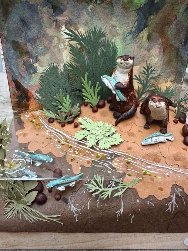 Diorama sculpture of 2 brown otters, one holds a fish in its hand and the other stands near a beached fish. They stand on a dirt shore with fish swimming nearby and cut greenery all around.