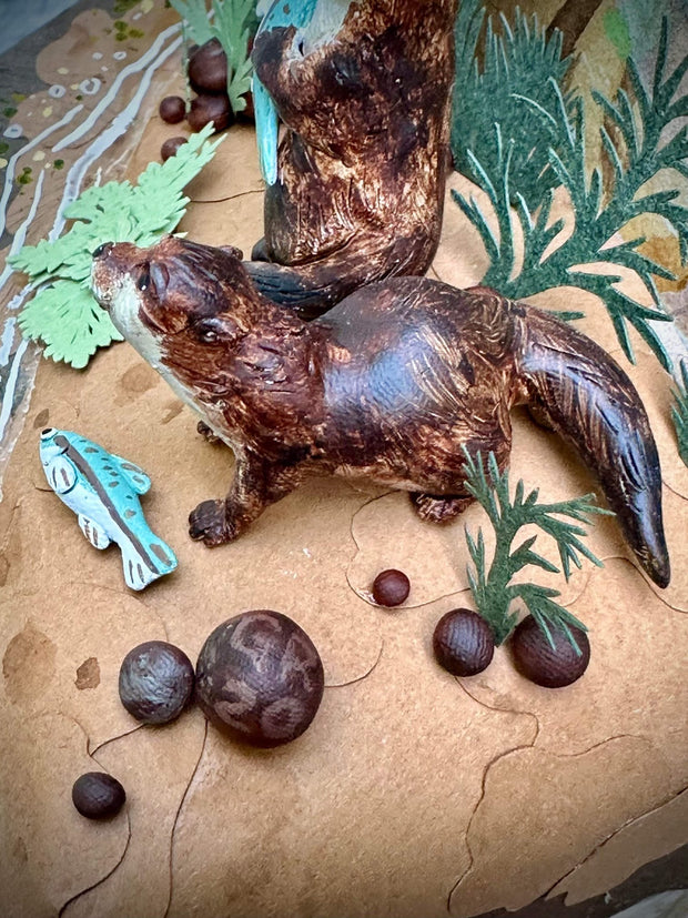 Diorama sculpture of 2 brown otters, one holds a fish in its hand and the other stands near a beached fish. They stand on a dirt shore with fish swimming nearby and cut greenery all around.